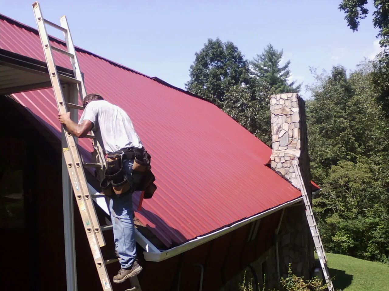 Roofer on a ladder beside a red metal roof and stone chimney — Soffit & Fascia Repair by Roofing Brothers in Cuddy